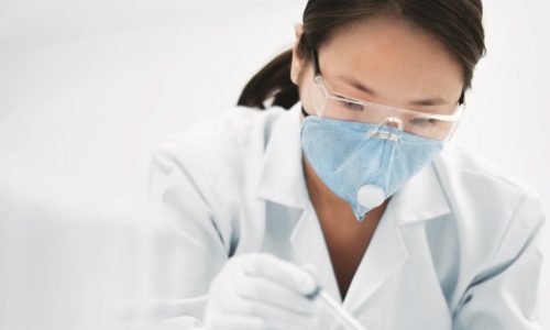 Asian woman scientist holding several test tubes and filling them with blue chemical by dropper, she is wearing safety goggle, glove, protective mask and lab coat.